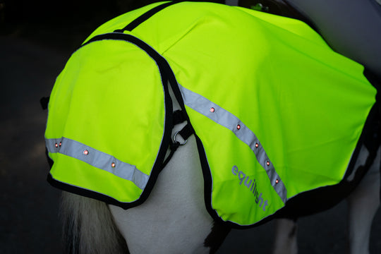 Horse wearing the EquiLight LED hi-vis exercise rug with integrated red LED safety lights and reflective trim. Rear view of the EquiLight LED exercise rug showing highway-code compliant red LED lights for rider and horse visibility. Close-up of the integrated USB-rechargeable red LED lighting built into the EquiLight hi-vis horse exercise rug. Breathable mesh fabric and reflective detailing used in the EquiLight LED hi-vis quarter sheet for horses. EquiLight LED hi-vis exercise rug laid flat showing shape, 